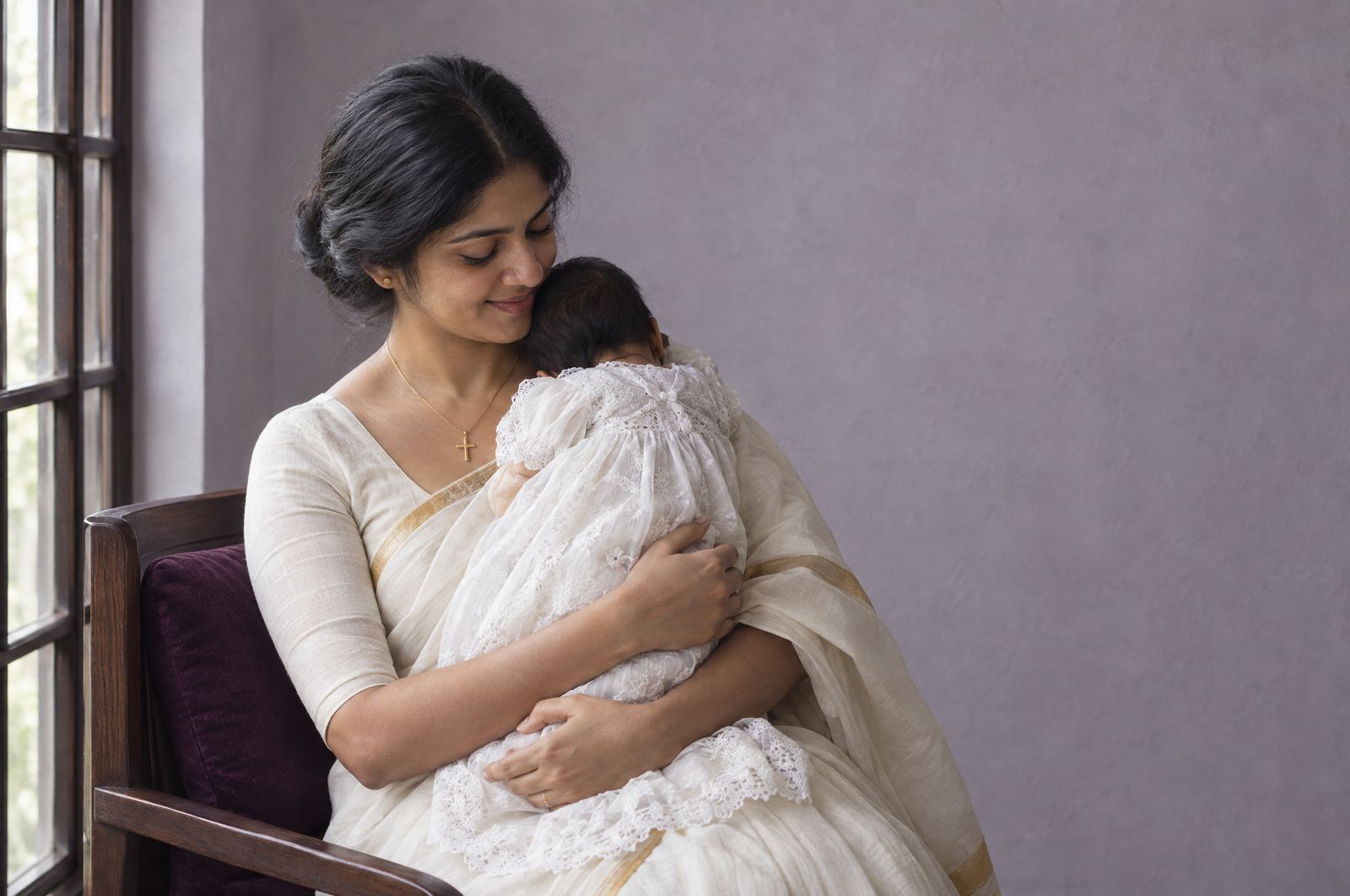 A mother holds her baby in a white christening gown by a window