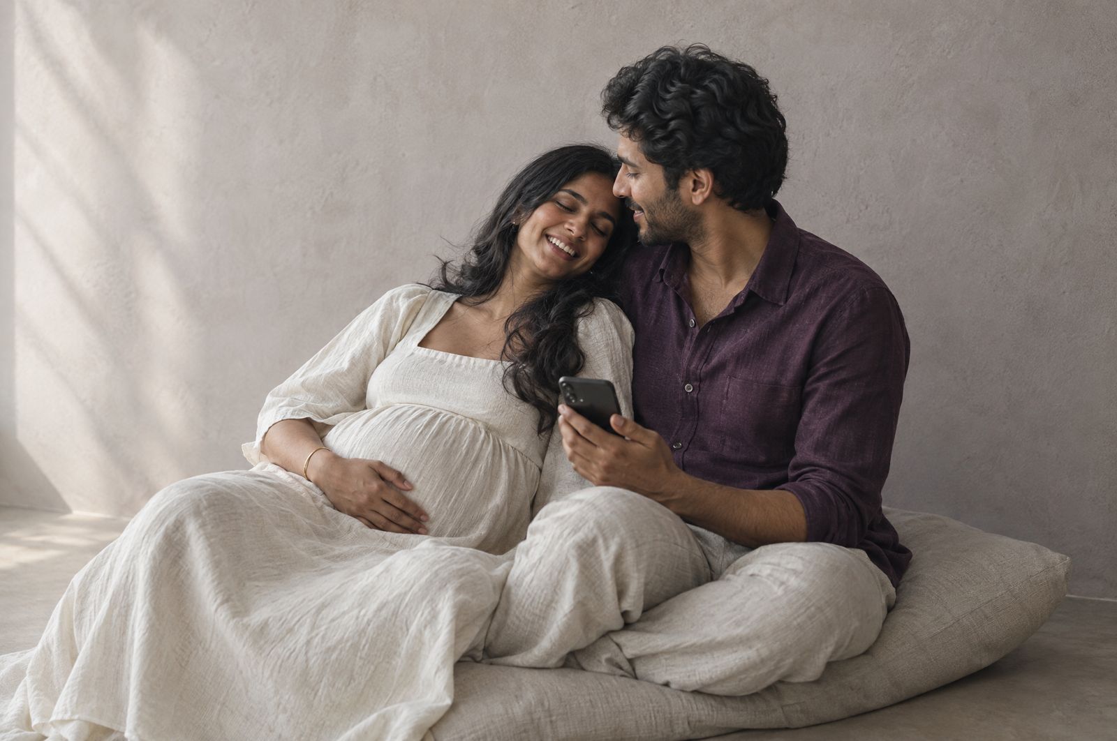 An expecting couple laugh together on a floor cushion, a phone between them