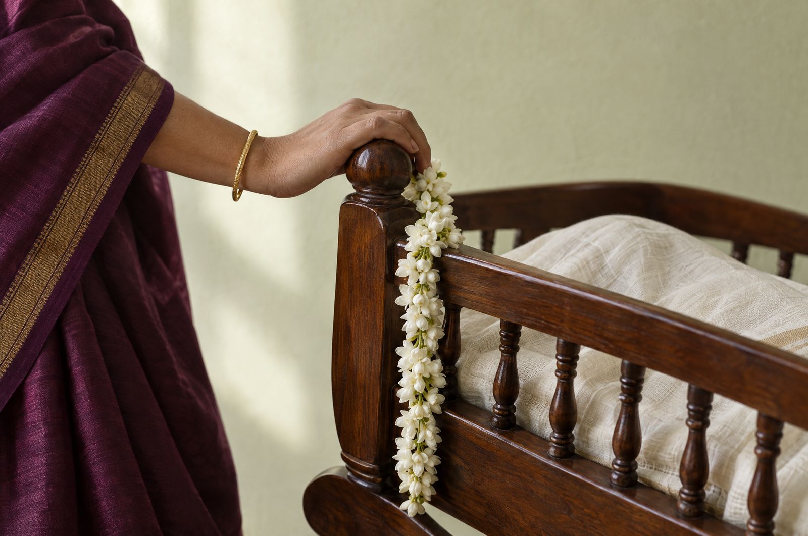 A mother's hand rests on a teak cradle tied with jasmine